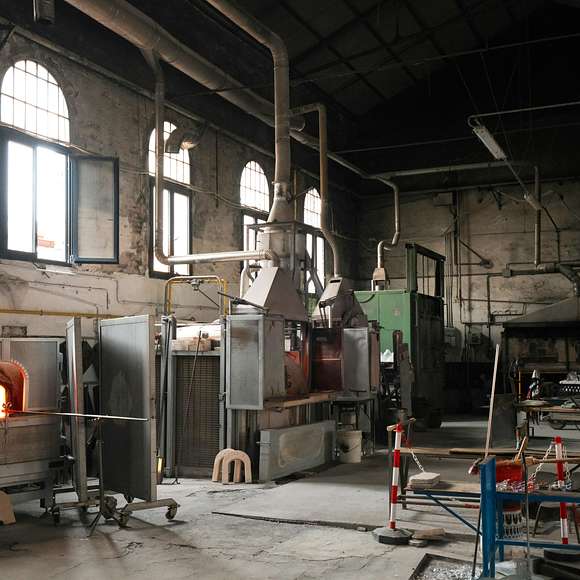 An interior view of a traditional glassblowing workshop with large furnaces, ventilation pipes, and scattered tools and materials throughout the space.