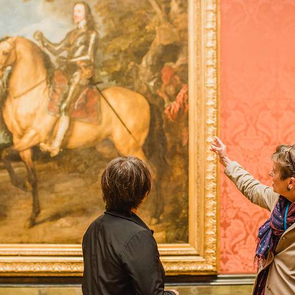 Tour guide gesturing toward a grand equestrian portrait while discussing it with a guest inside the National Gallery.