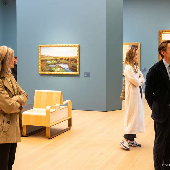 People admire framed landscape paintings in a brightly lit room with blue walls.