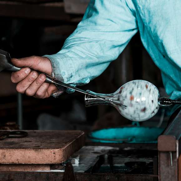 A glassblower in a light blue shirt shapes a speckled glass orb at the end of a blowpipe inside a workshop, using tools to refine its form.