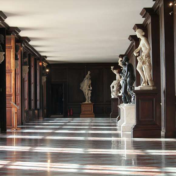 A long wood-paneled gallery inside Hampton Court Palace with marble statues and busts displayed along the walls, sunlight streaming across the checkered floor.