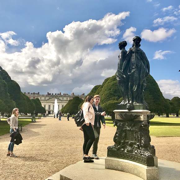 Visitors walking past a bronze statue of two figures in the gardens of Hampton Court Palace, with manicured trees and the palace fountain in the background.