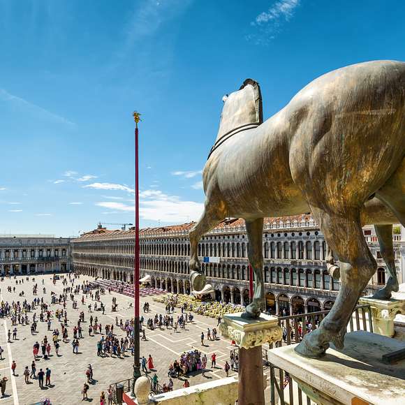 View of Piazza San Marco from the basilica terrace, framed by bronze horse statues and red flagpoles.