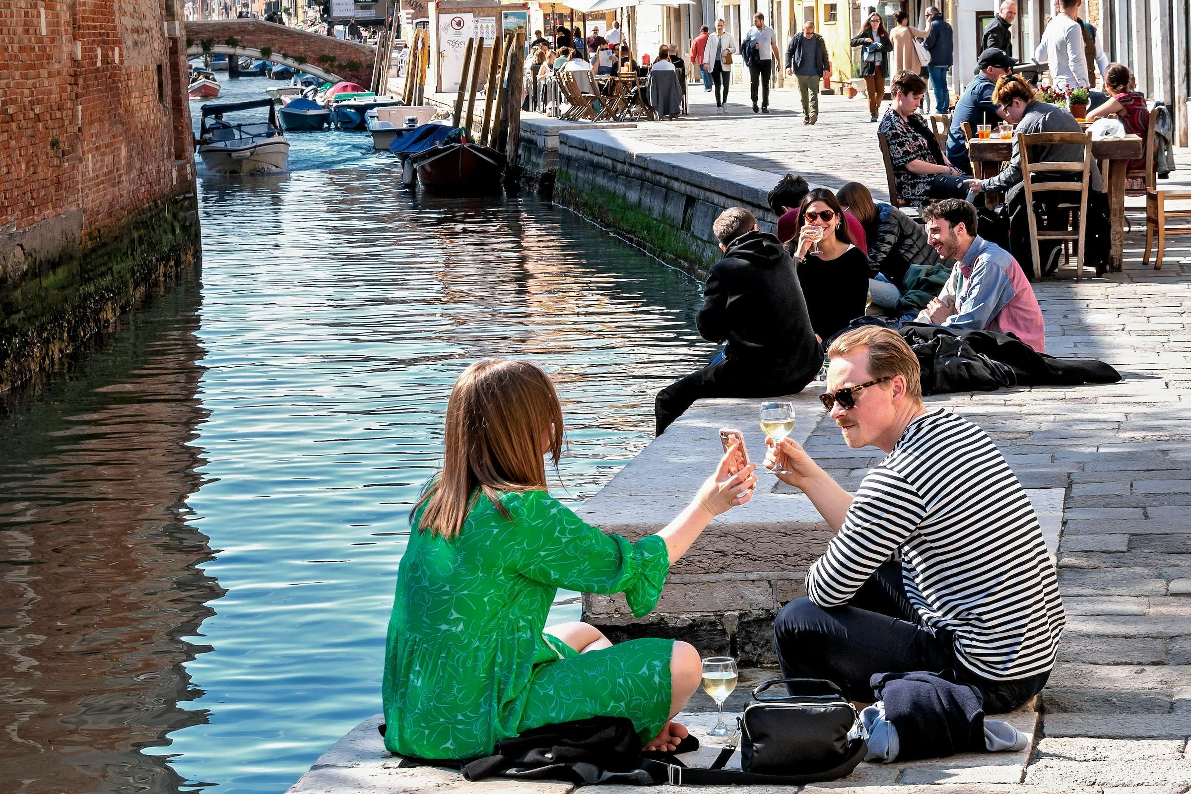 People relax with drinks along a quiet canal in Cannaregio on a sunny afternoon.