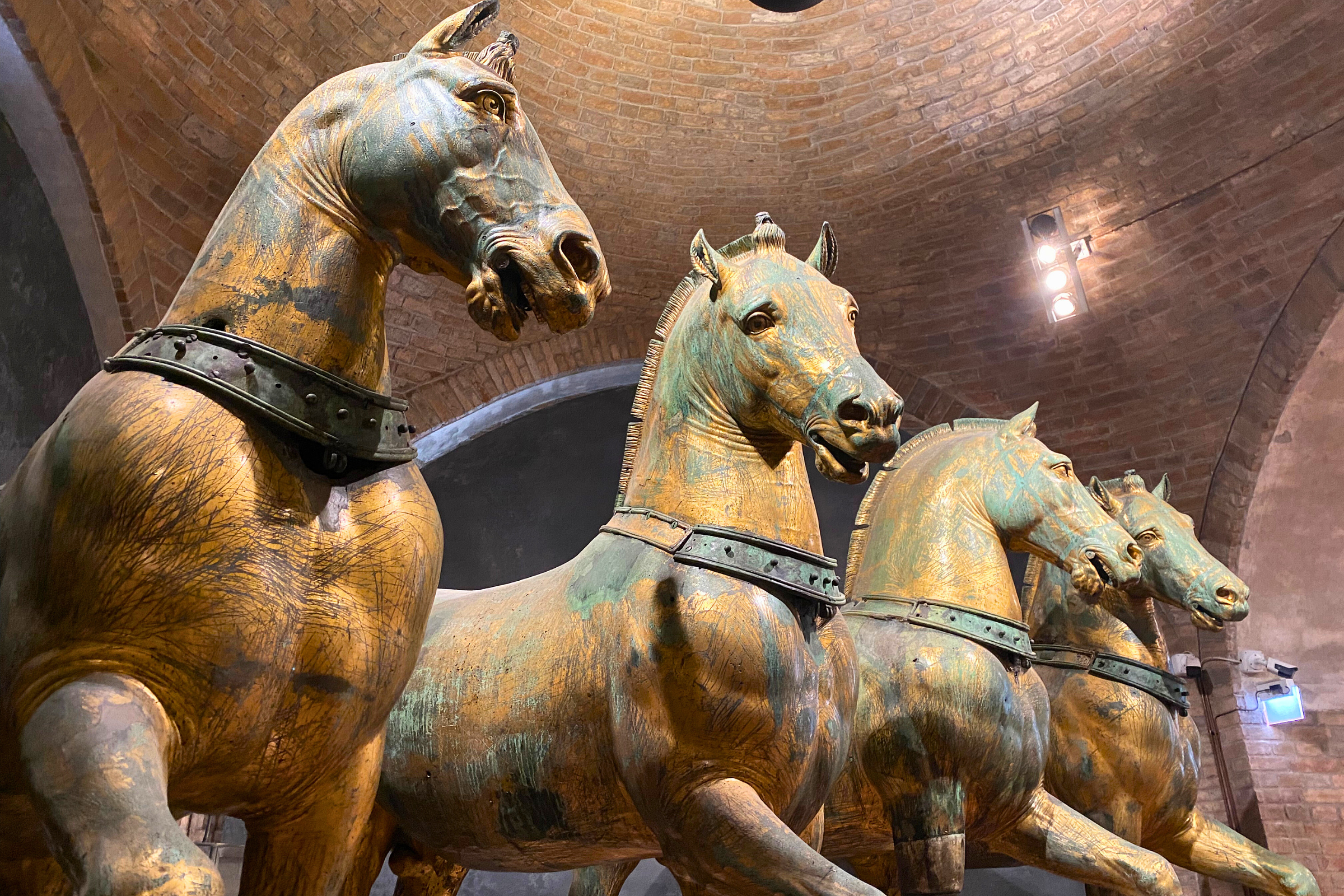 Ancient bronze Horses of Saint Mark inside the basilica, set against a vaulted brick ceiling and museum lighting.