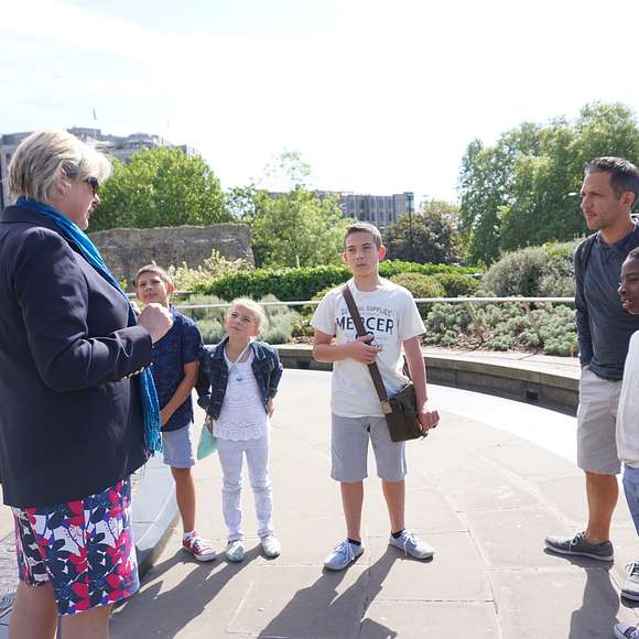 Tour guide engaging a group of children and an adult outside near the Tower of London ruins, with London Underground signage visible.