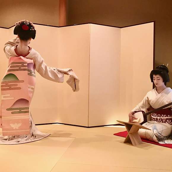 A geisha in a white kimono performs a traditional dance while another sits nearby playing a shamisen, both set against a minimalist folding screen in a tatami-mat room.